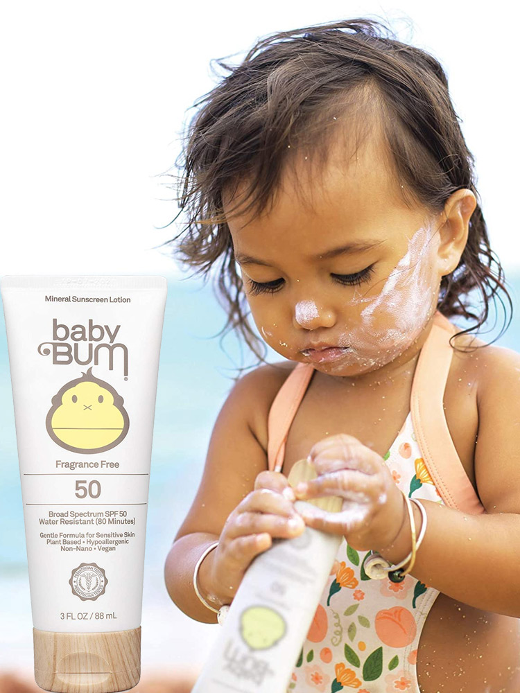 a baby girl on the beach holding a tube of baby bum sunscreen