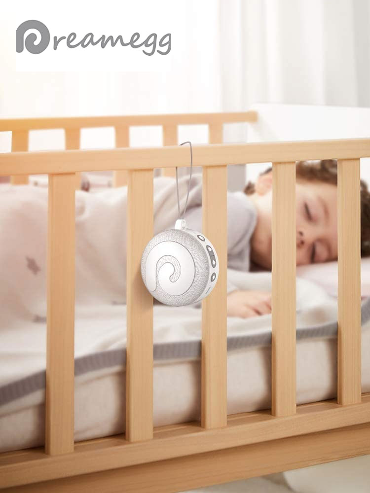 the dreamegg portable white noise machine hanging on the side of a crib with a baby sleeping inside