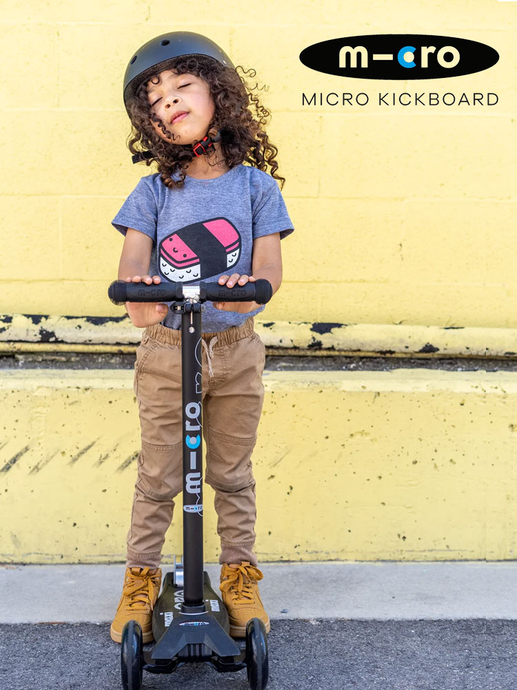 a boy standing on a micro maxi deluxe scooter against a yellow wall