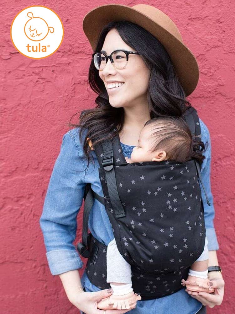 woman with glasses carrying a baby in a tula free-to-grow baby carrier against a red wall