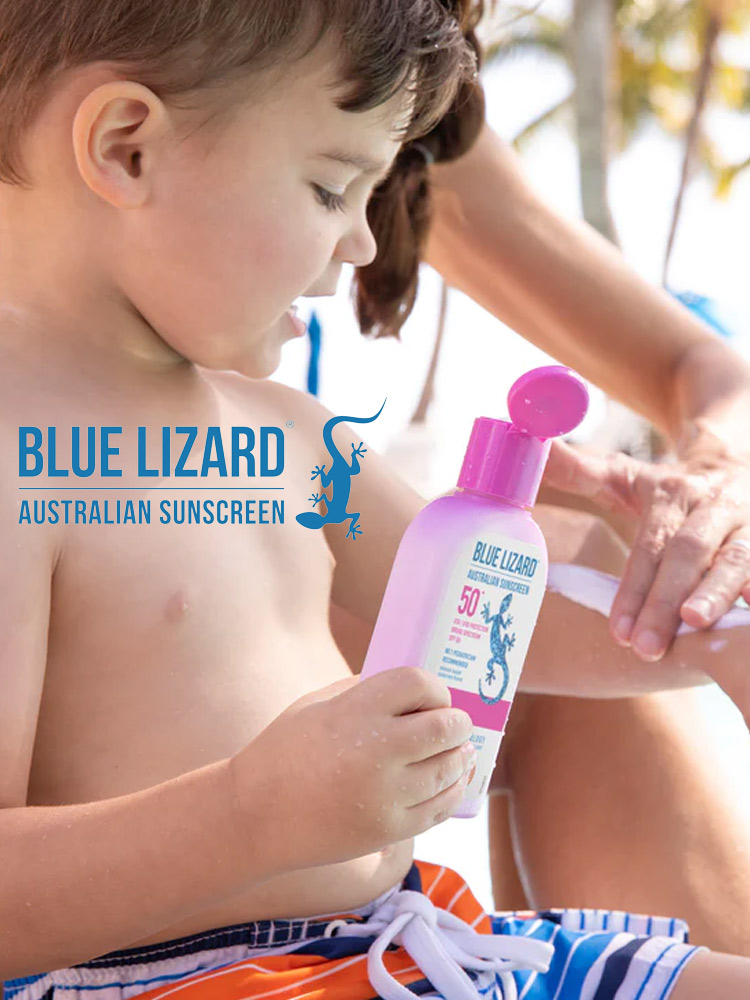 a young boy on the beach holding a bottle of blue lizard sunscreen while a parent applies sunscreen to his arm