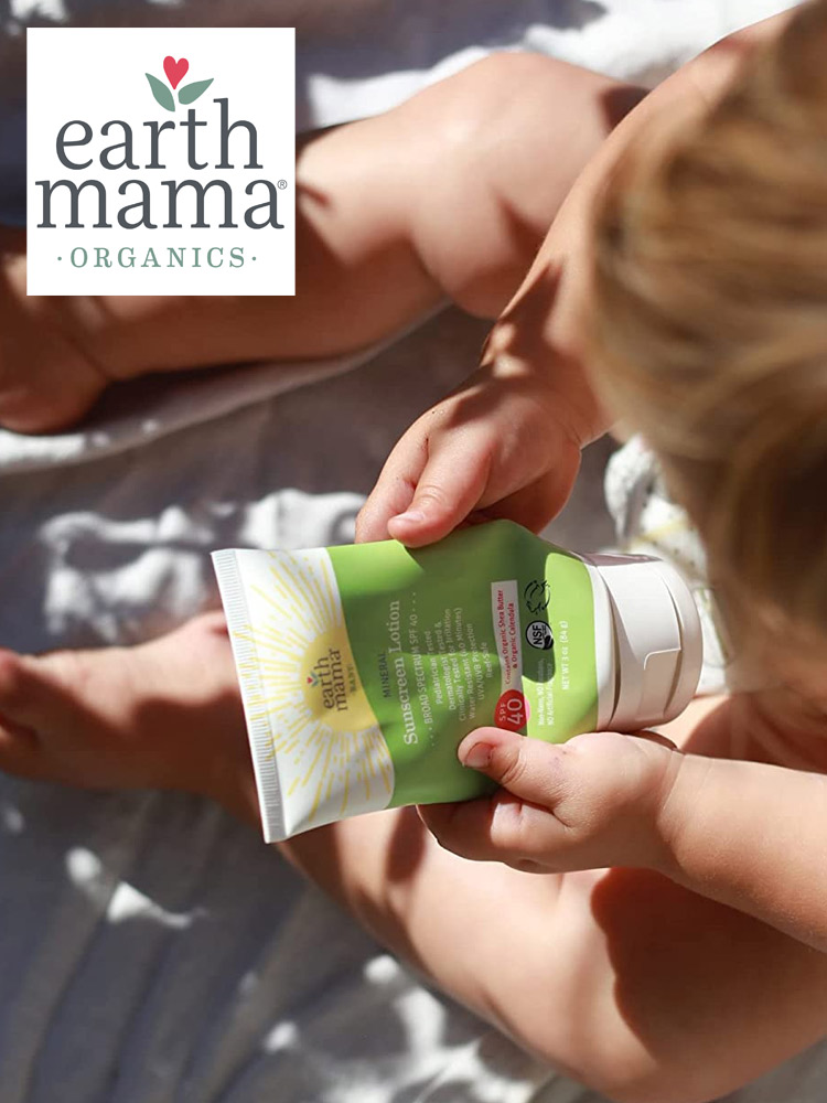 a baby sitting on the beach holding a tube of earth mama sunscreen