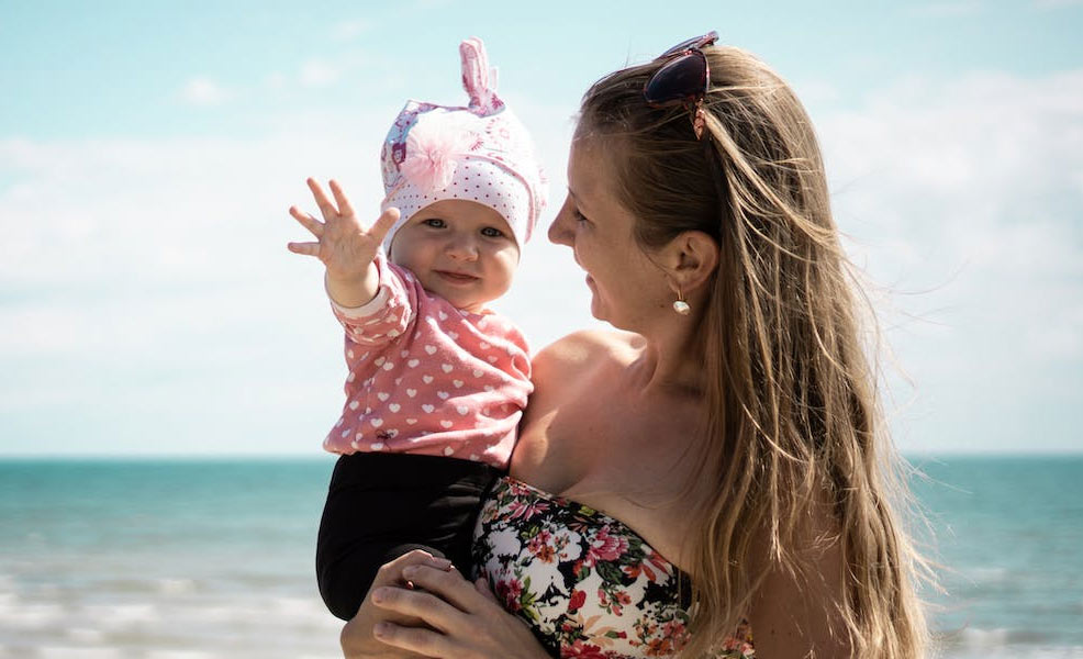 a mom carrying a baby on beach wearing sunscreen and a hat