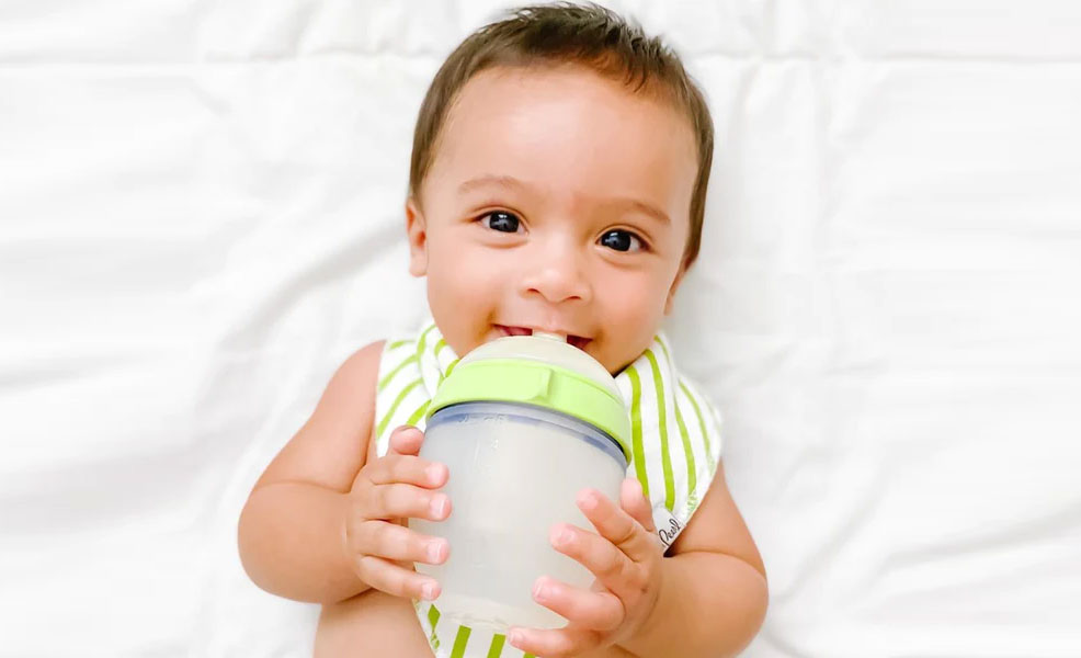 baby boy laying down with a striped bib and drinking from a comotomo baby bottle