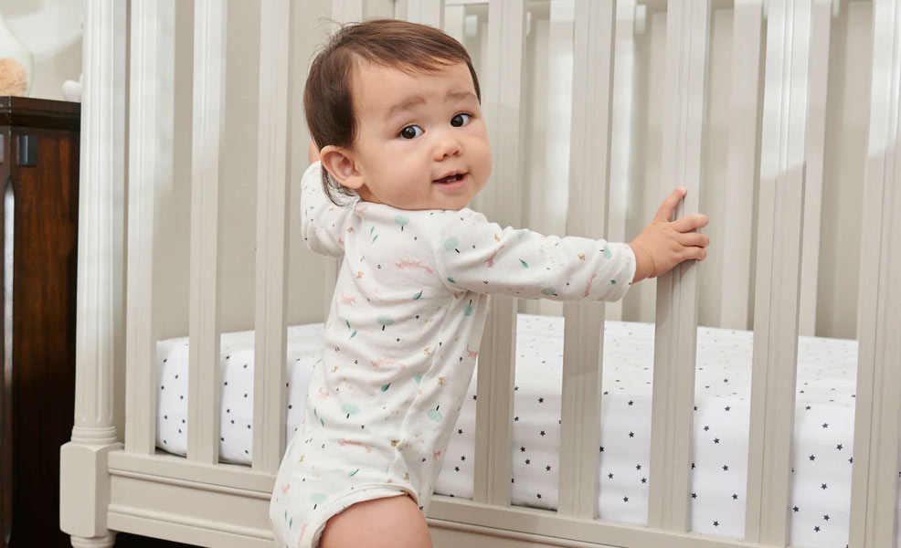 a baby smiling while holding onto the bars of a crib that has the moonlight slumber crib mattress inside