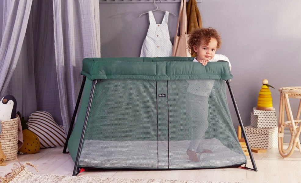 a nursery with a green travel crib with a toddler standing inside and looking at the camera