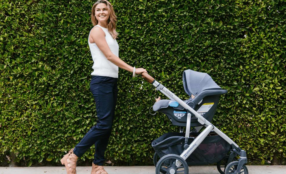 mom pushing the uppababy vista travel system with attached infant car seat against background of bushes