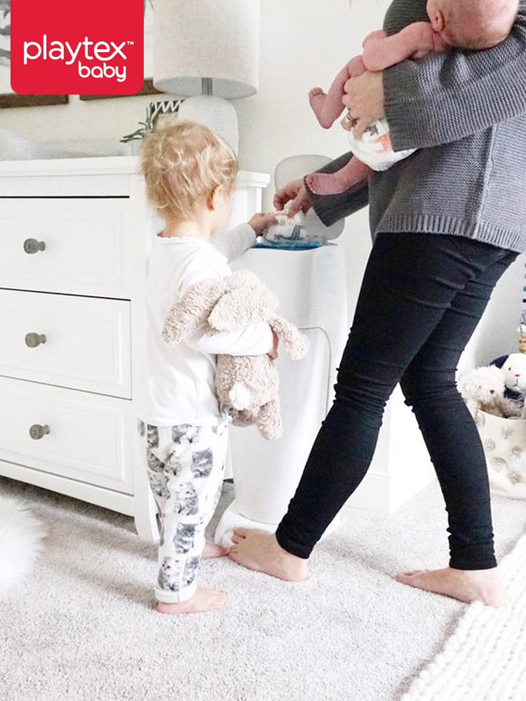 a mom holding a baby while a toddler helps her dispose of a dirty diaper in the playtex genie diaper pail