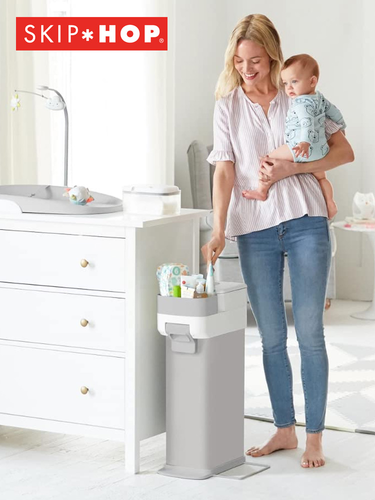 a mom holding a baby in a nursery and using the skip hop diaper pail