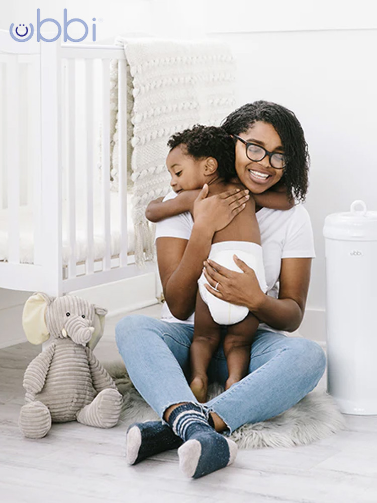 a mom hugging a toddler boy in a diaper while sitting next to the ubbi steel diaper pail